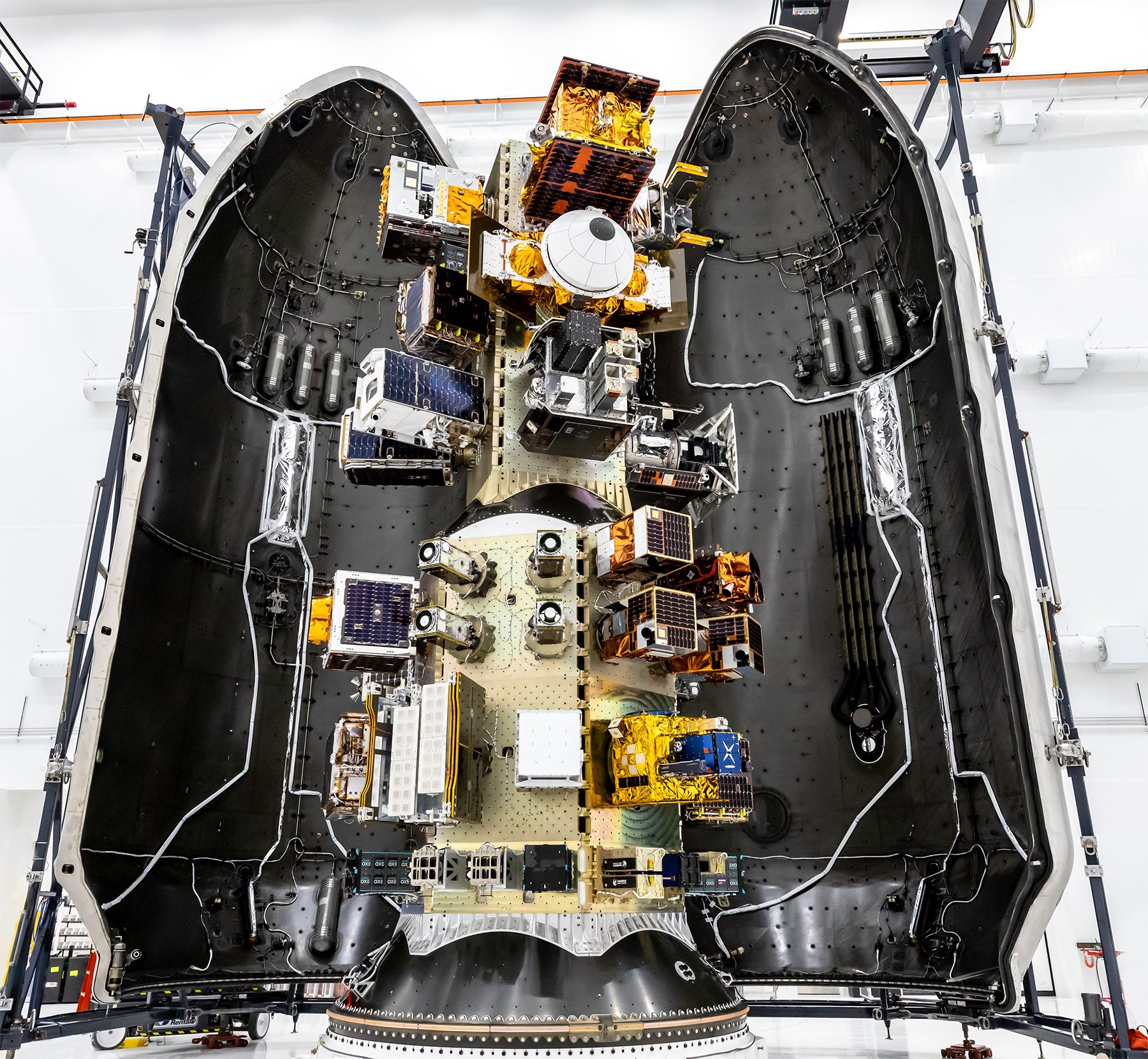 Top‑down view of a SpaceX payload fairing interior showing mounted satellites and hardware arranged along the center structure, surrounded by the curved black fairing walls.